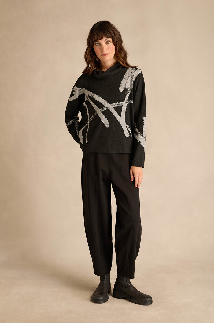 A woman stands against a beige background wearing the Paint Stripes Long Sleeve Top—a black relaxed-fit sweater with gray abstract lines—paired with black loose pants and black boots.