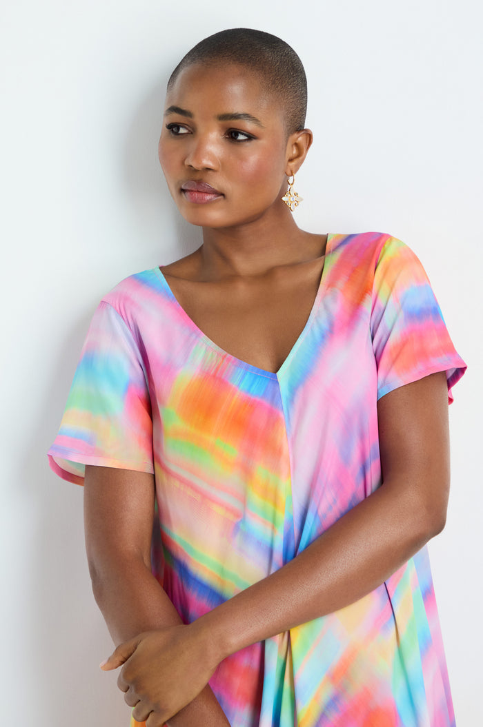 A person with short hair models the Painted Horizon Bubble Dress, featuring a colorful tie-dye design, while standing against a white background and looking slightly to the side.