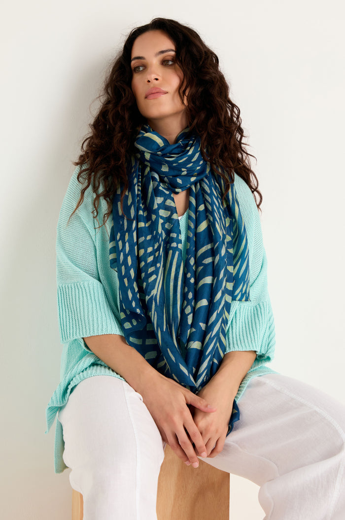 A woman with long curly hair wears a light blue sweater, white pants, and the Painted Waves Silk Scarf, sitting on a wooden stool against a plain light background.