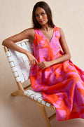 A woman sits on a white woven chair, looking at the camera while wearing the Palm Mirage Linen Flared Dress—a sleeveless fit-and-flare linen dress in pink and orange patterns.