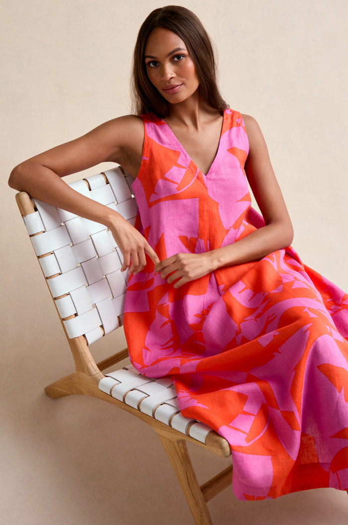 A woman sits on a white woven chair, looking at the camera while wearing the Palm Mirage Linen Flared Dress—a sleeveless fit-and-flare linen dress in pink and orange patterns.