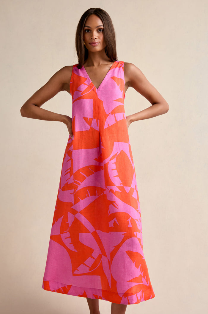 A woman models the Palm Mirage Linen Flared Dress, a sleeveless, ankle-length fit-and-flare style featuring a pink and orange abstract leaf pattern, set against a plain beige background.