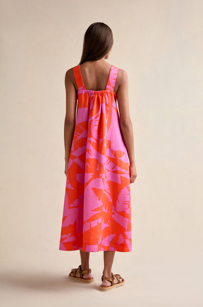 A woman faces away, wearing the Palm Mirage Linen Flared Dress—sleeveless, ankle-length with a pink and orange palm print—paired with flat brown sandals against a plain background.