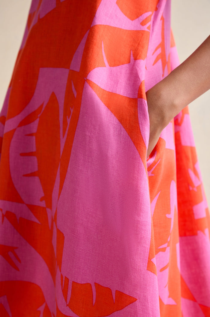 A person’s hand rests in the pocket of the Palm Mirage Linen Flared Dress, showcasing its bold pink and orange abstract leaf pattern.