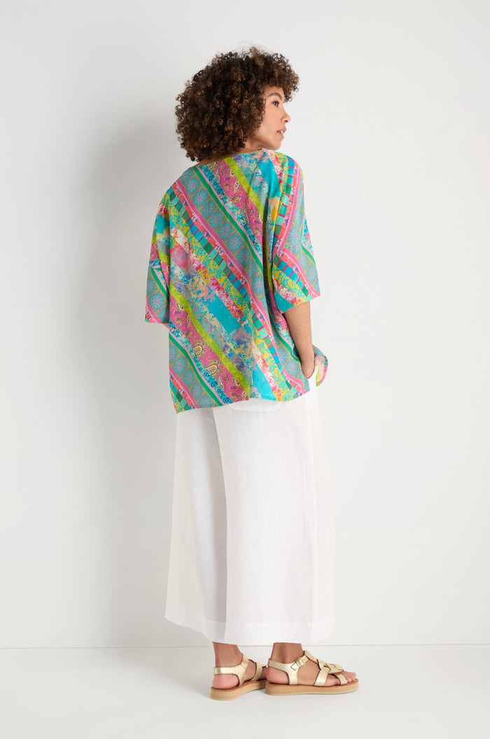 A person stands sideways in a Patchwork Stripe Boxy Top, wide white pants, and beige sandals against a plain white background.