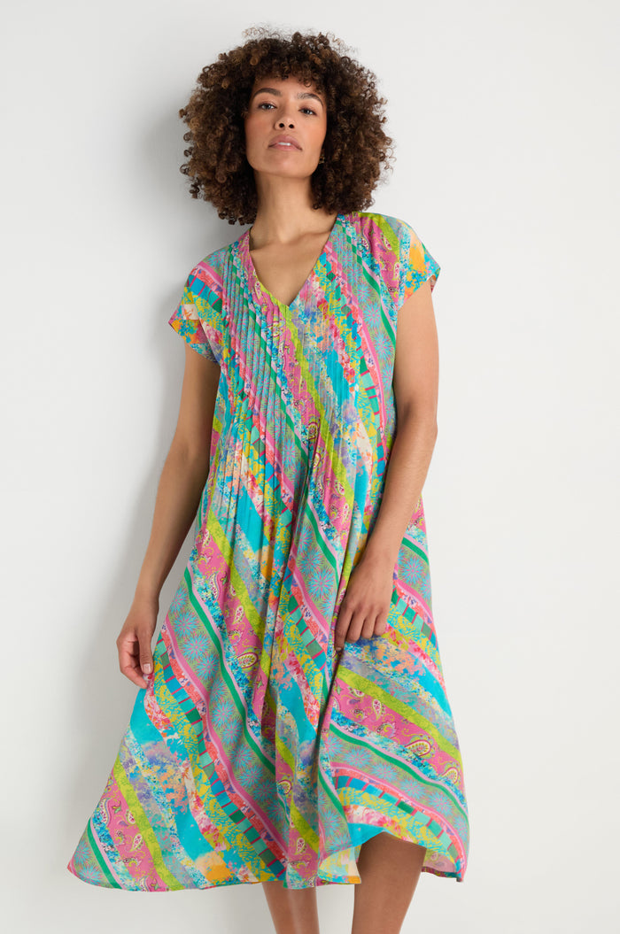 A woman stands against a plain background wearing the Patchwork Stripe Pleated Dress, featuring colorful patterns, short sleeves, a V-neck, pleats, and a flattering fit-and-flare silhouette.