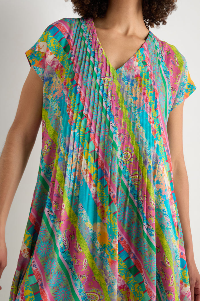 A person wears the Patchwork Stripe Pleated Dress, featuring colorful diagonal stripes and abstract patterns in pink, green, blue, and yellow with pleats, neckline detailing, and a flattering fit-and-flare silhouette.