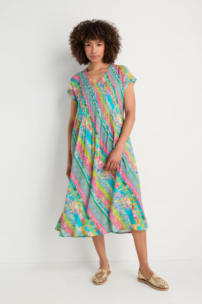 A person models the Patchwork Stripe Pleated Dress—loose, knee-length with short sleeves—paired with gold woven slip-on shoes, standing against a plain white background.