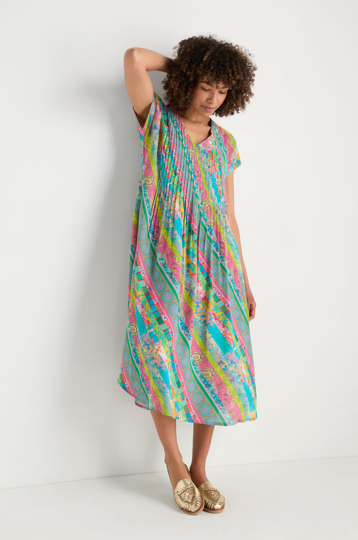 A person stands against a plain white wall, wearing the Patchwork Stripe Pleated Dress with short sleeves and gold slip-on shoes.