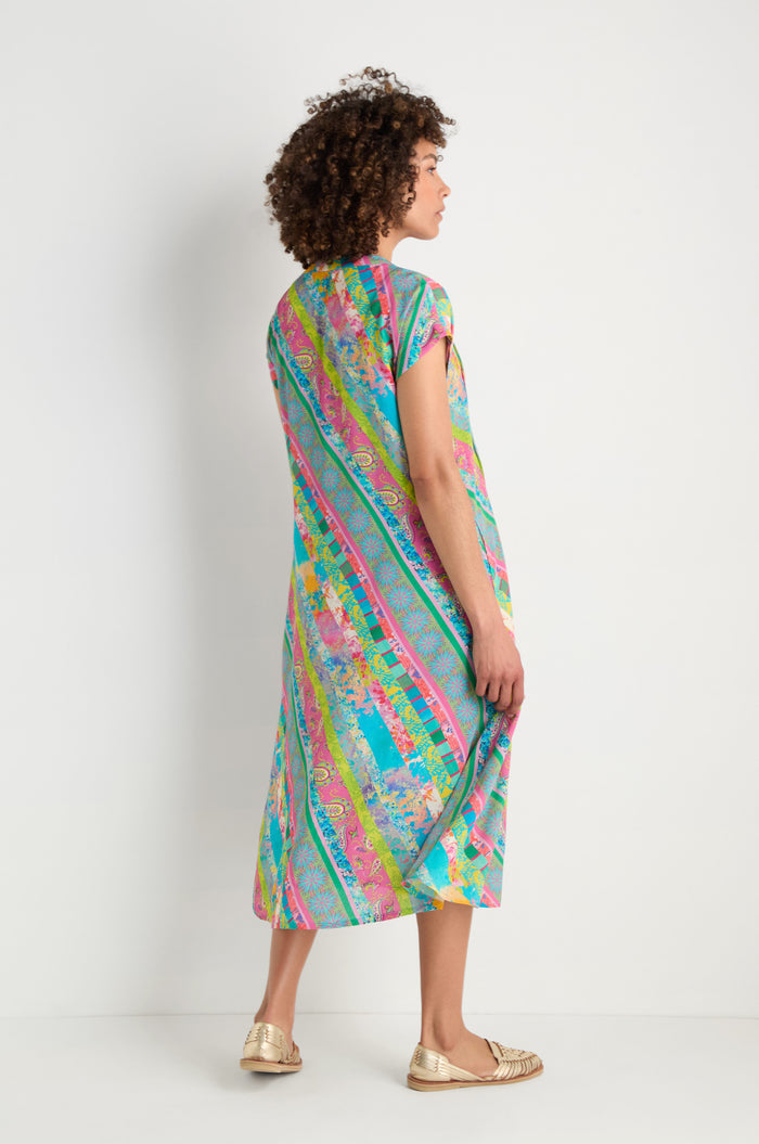 A person stands against a white backdrop, facing away, wearing the Patchwork Stripe Pleated Dress with a fit-and-flare silhouette and gold flats.