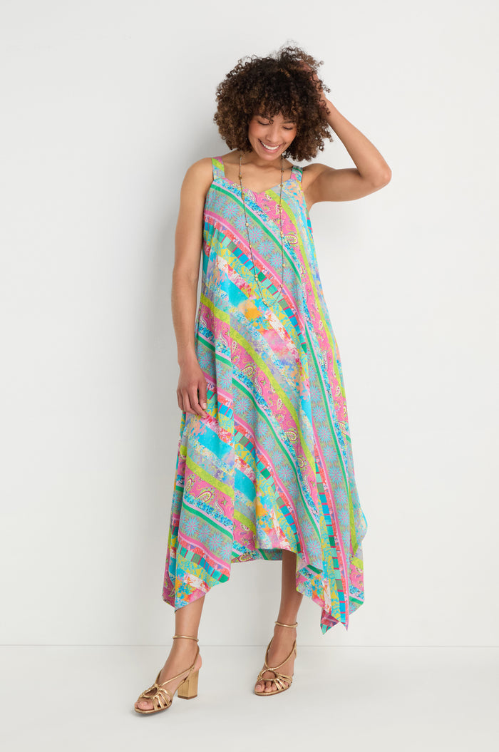 A woman stands against a white wall wearing the Patchwork Stripe Summer Dress with an asymmetric hem and gold sandals.