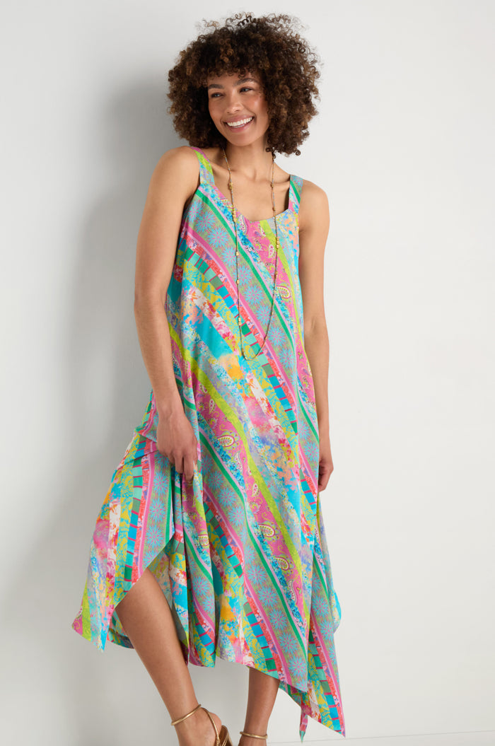 A woman smiles as she stands against a plain light wall, wearing the Patchwork Stripe Summer Dress—a colorful, sleeveless viscose crepe dress featuring diagonal stripes and a floral pattern.