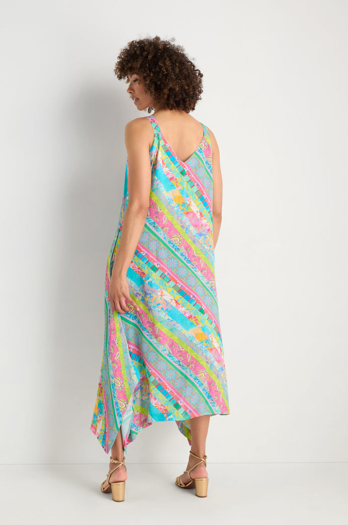 A woman faces away, wearing the Patchwork Stripe Summer Dress with an asymmetrical hem and sandals. The vibrant viscose crepe fabric drapes elegantly, making it perfect for warm days.
