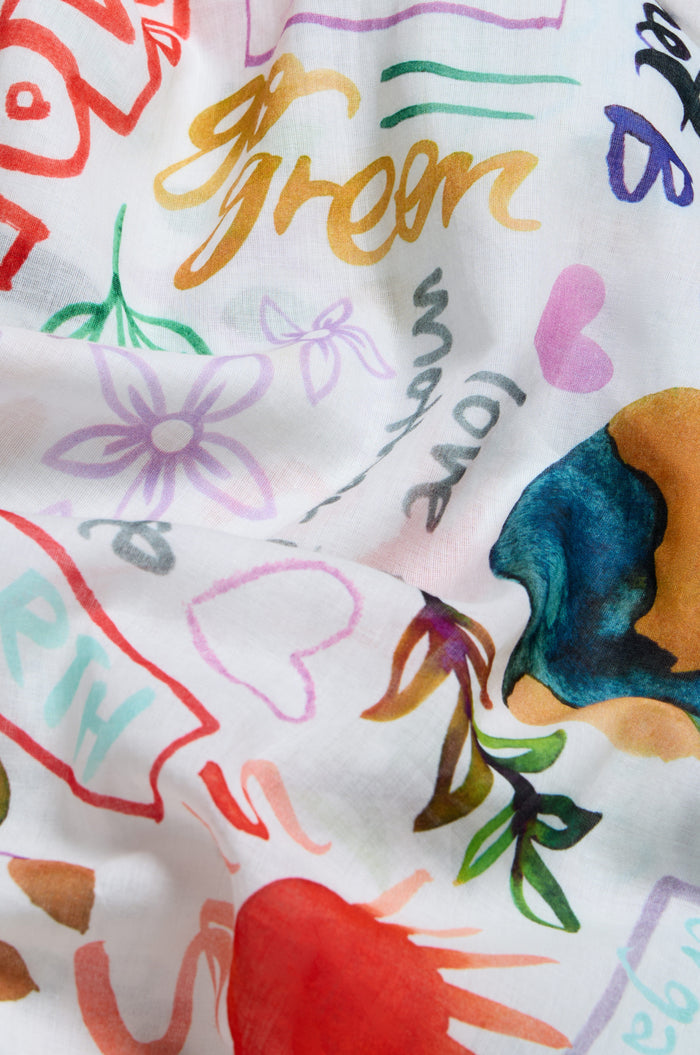 The Peace and Love Cotton Scarf is a lightweight white scarf with vibrant floral prints, colorful doodles, hearts, abstract shapes, and playful words like "green," "love," and "earth.