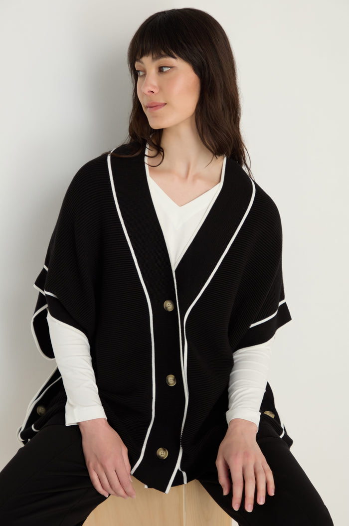 A woman wears the Piped Detail Cardigan with white trim and gold buttons over a white shirt, seated on a light wooden stool against a plain background.