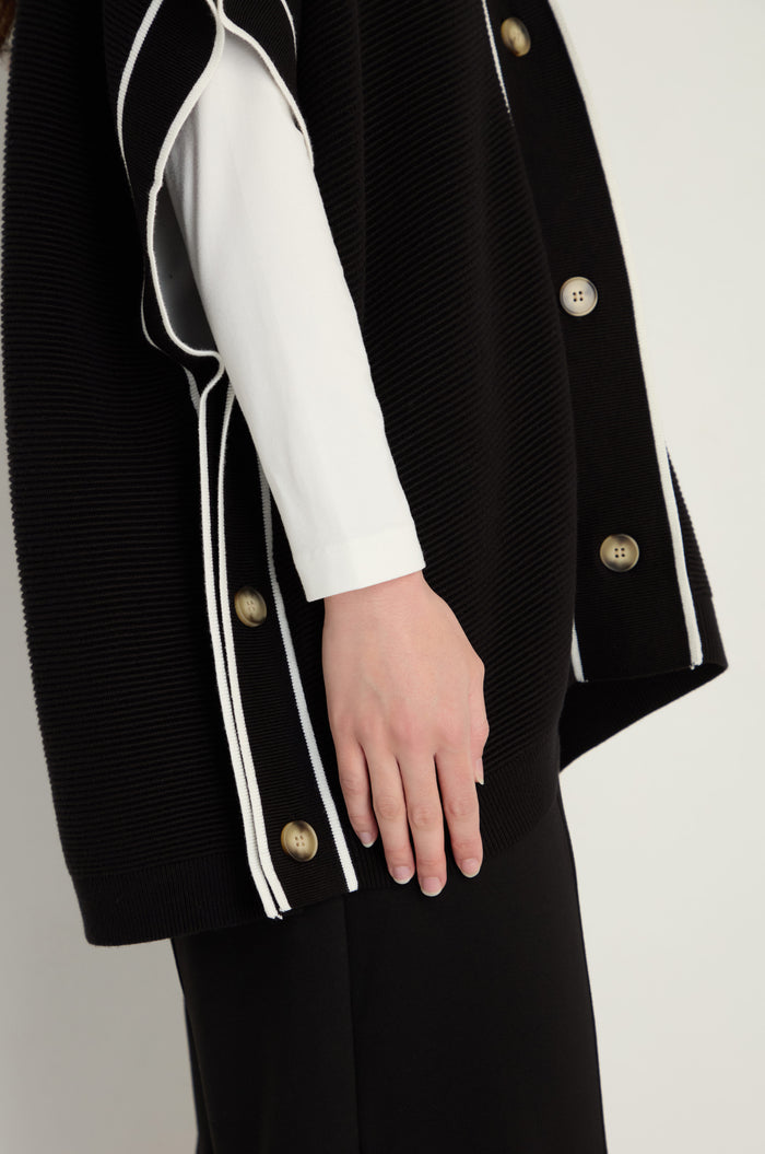 A person wears the Piped Detail Cardigan, featuring gold buttons and white trim, layered over a white long-sleeve top and shown from shoulders to mid-thigh.