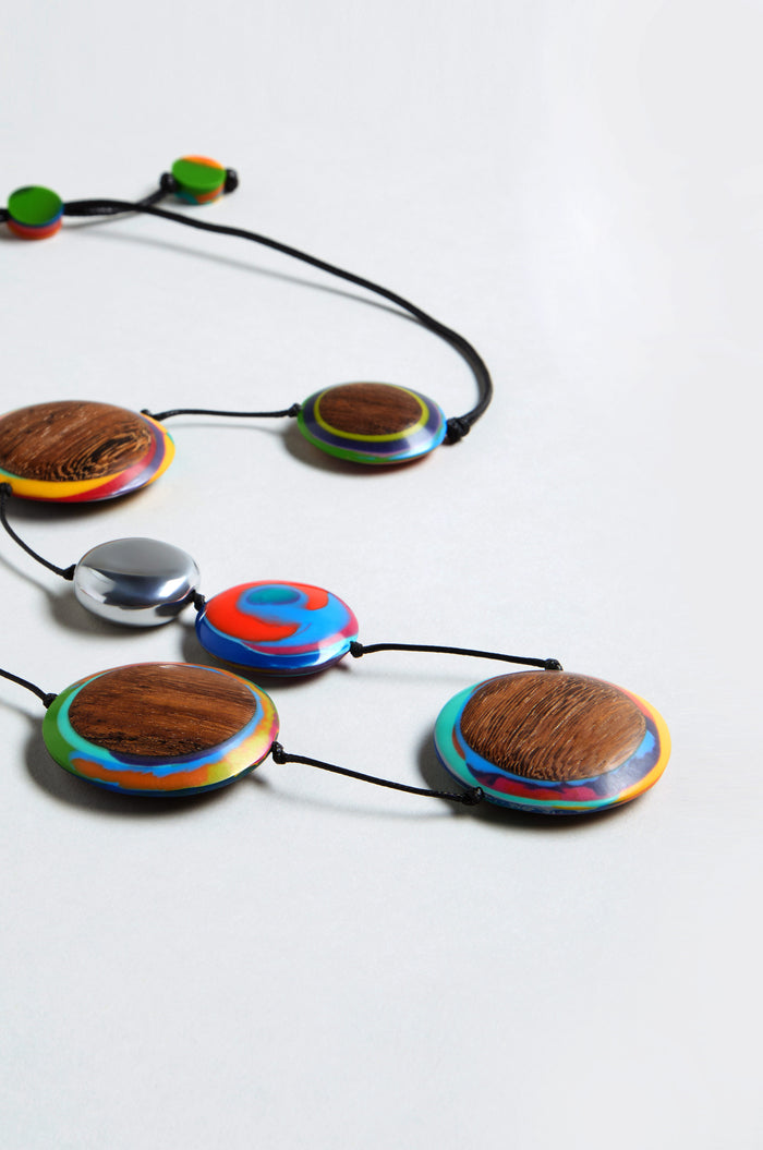 The Planetary Necklace features large, round beads with colorful patterns and wood textures, plus a metallic recycled aluminium disc, all on a black cord and displayed on a white surface.