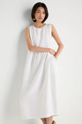 A woman stands against a white background wearing the Pleat Front Linen Bubble Dress, an ankle-length, sleeveless linen dress with textured fabric and gold earrings, with one hand in her pocket.