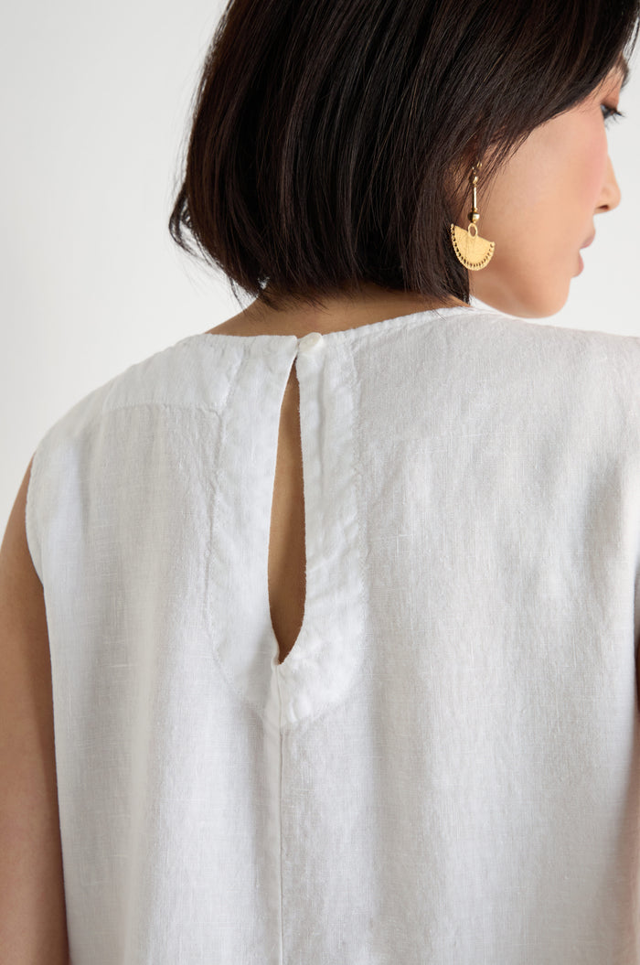 A woman with short dark hair, seen from behind, wears a sleeveless white Pleat Front Linen Bubble Dress by Grizas with a back keyhole opening and gold half-moon earrings.
