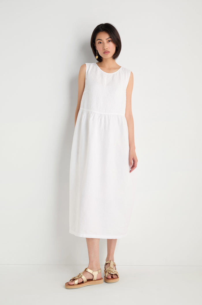 A woman with short dark hair and a neutral expression stands against a white wall, wearing the sleeveless, ankle-length Pleat Front Linen Bubble Dress and beige sandals.