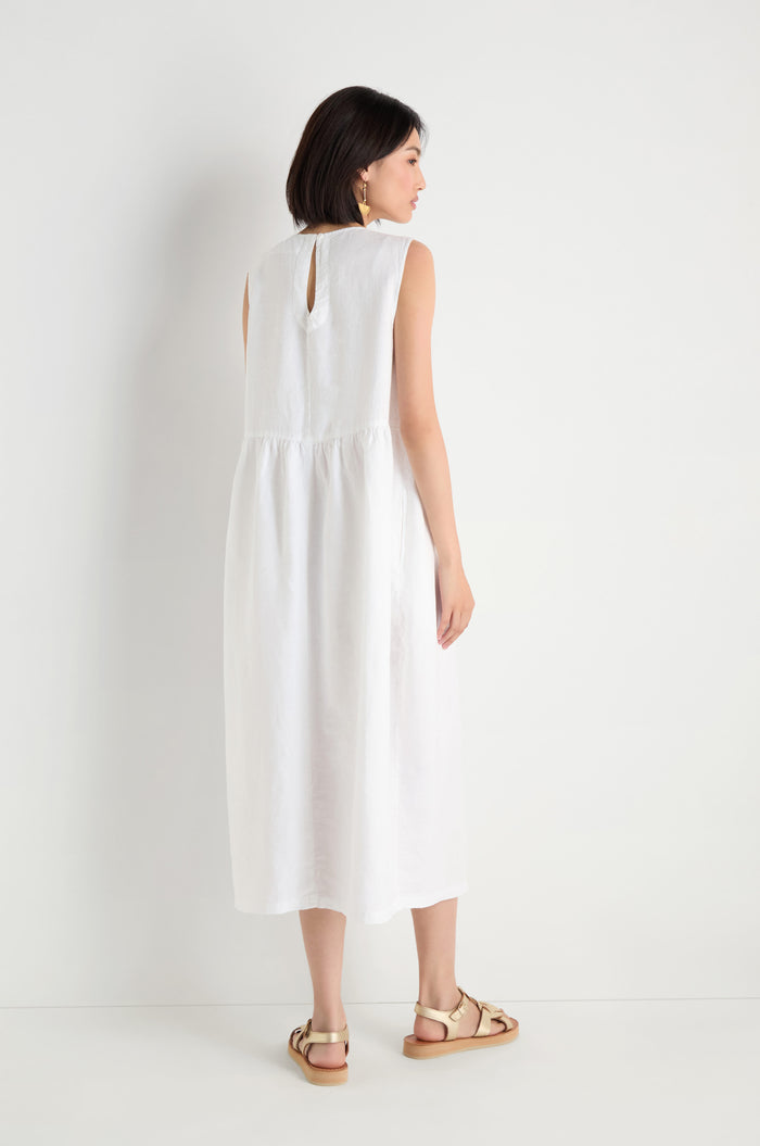 Woman standing against a white wall, facing away, wearing a sleeveless Pleat Front Linen Bubble Dress and beige sandals.