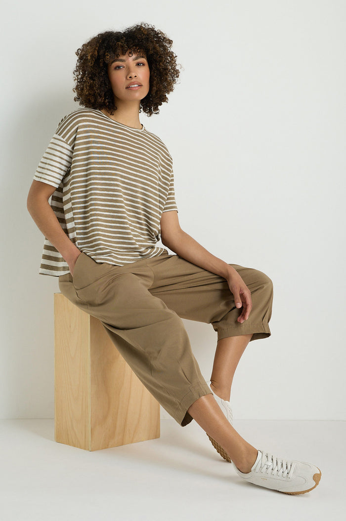 A person wearing a striped short-sleeve shirt, Pleated Jersey Trousers with an elasticated waist, and light sneakers sits on a wooden block against a plain white background.