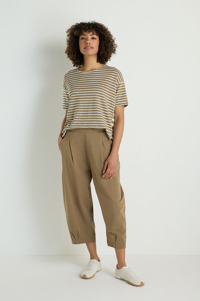 Person standing against a plain white wall, wearing a striped short-sleeve shirt, Pleated Jersey Trousers with an elasticated waist, and white sneakers, hands in pockets.