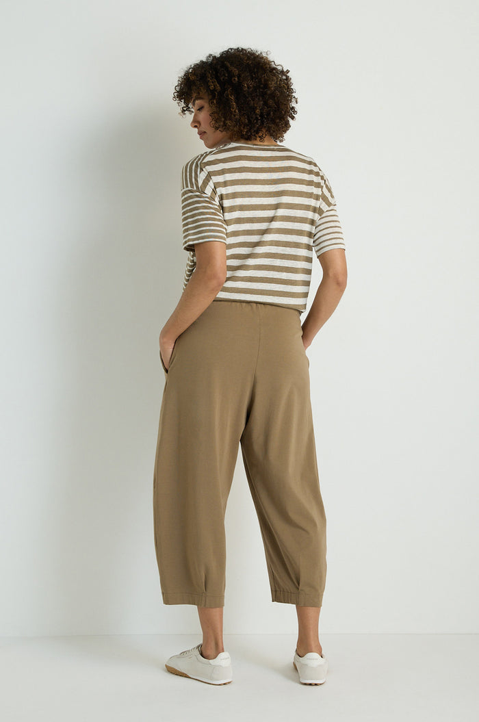 A person stands against a plain background, wearing Pleated Jersey Trousers with an elasticated waist, a striped t-shirt, and white sneakers. Facing away from the camera, their hands are tucked into their pockets.