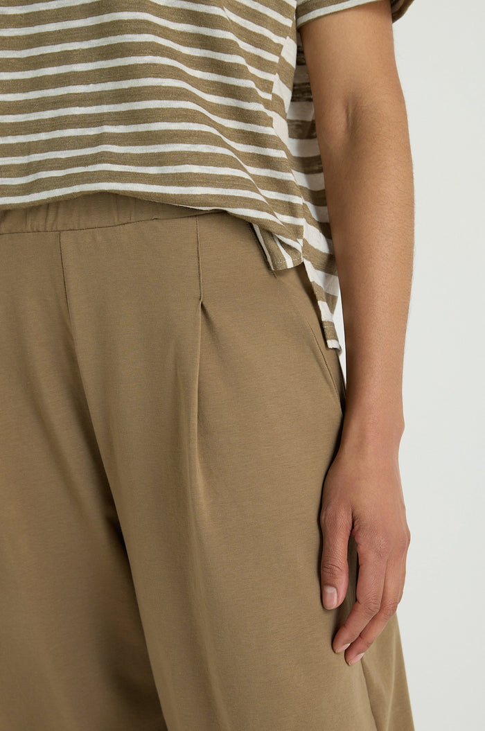 A person wears a beige and white striped shirt paired with Pleated Jersey Trousers featuring an elasticated waist, their right hand resting by their side.