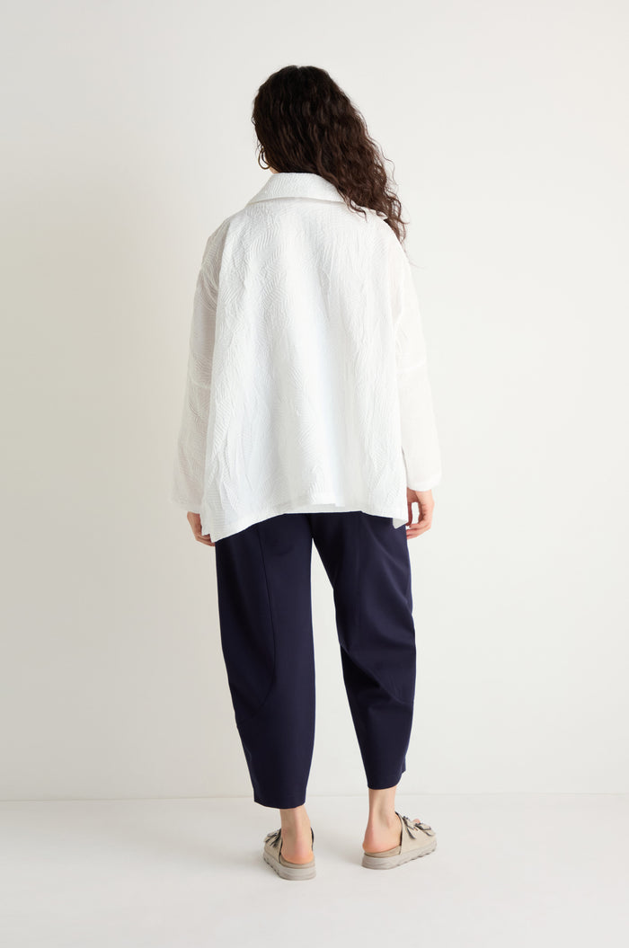 A person stands with their back turned, wearing the Pleated Leaf Short Jacket in white, paired with navy pants and beige slip-on shoes, pictured against a plain white background.