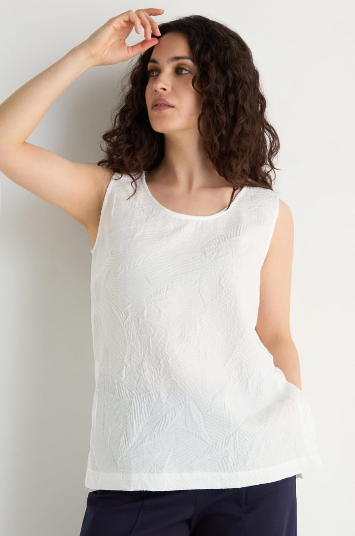 A woman with curly hair models the Pleated Leaf Top, a sleeveless white pleated top, paired with dark pants against a plain light background.