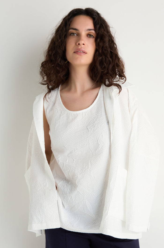 A woman with wavy brown hair wears the Pleated Leaf Top and a matching white jacket, both made from three-dimensional pleated fabric, standing against a plain light background.