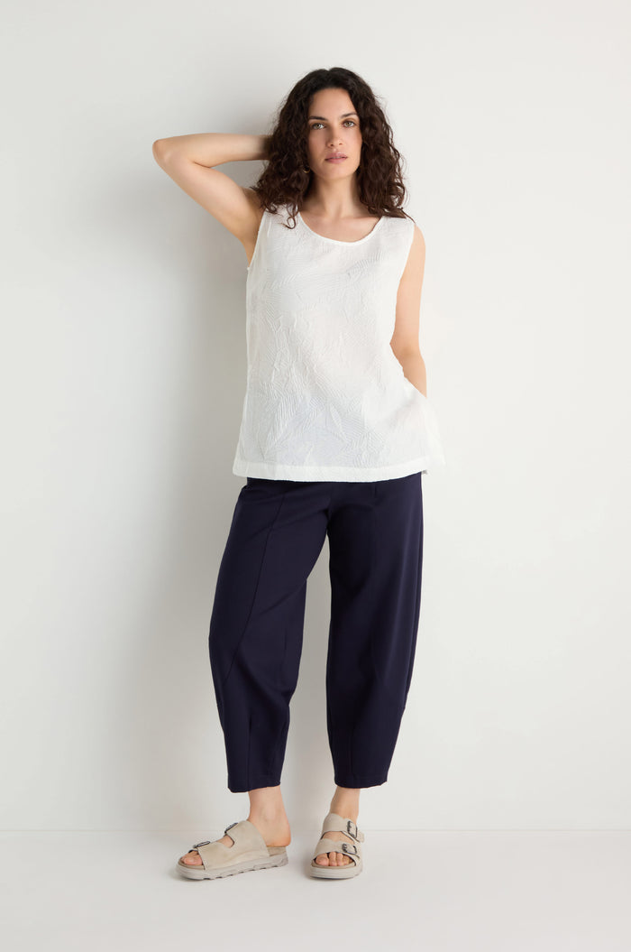A woman stands against a white wall wearing the Pleated Leaf Top, navy loose-fitting pants, and beige sandals, with one hand behind her head.