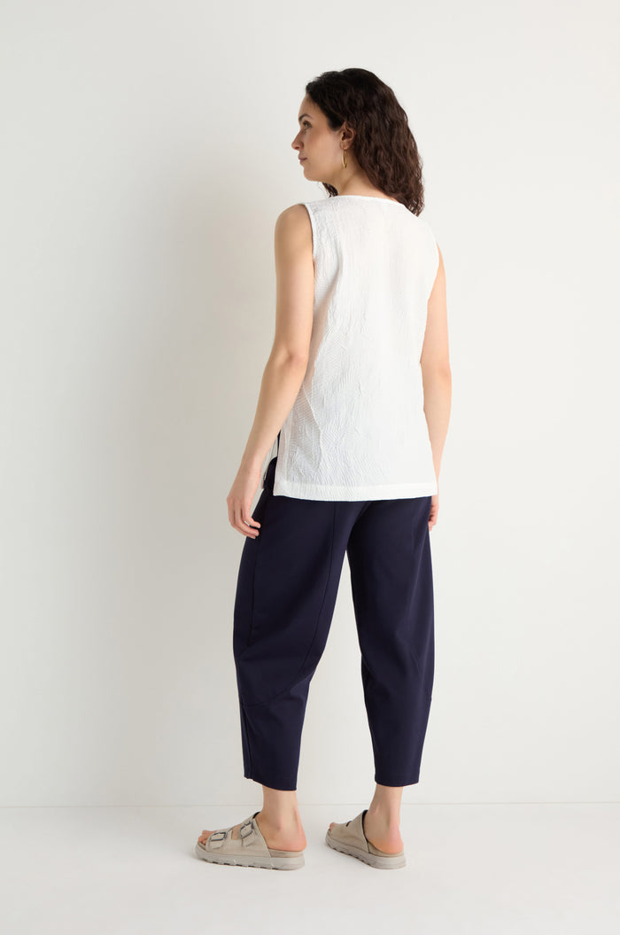 A person stands against a white wall, facing away, wearing the Pleated Leaf Top. The raised leaf pattern and three-dimensional pleated fabric are highlighted as they pair it with navy trousers and beige sandals.
