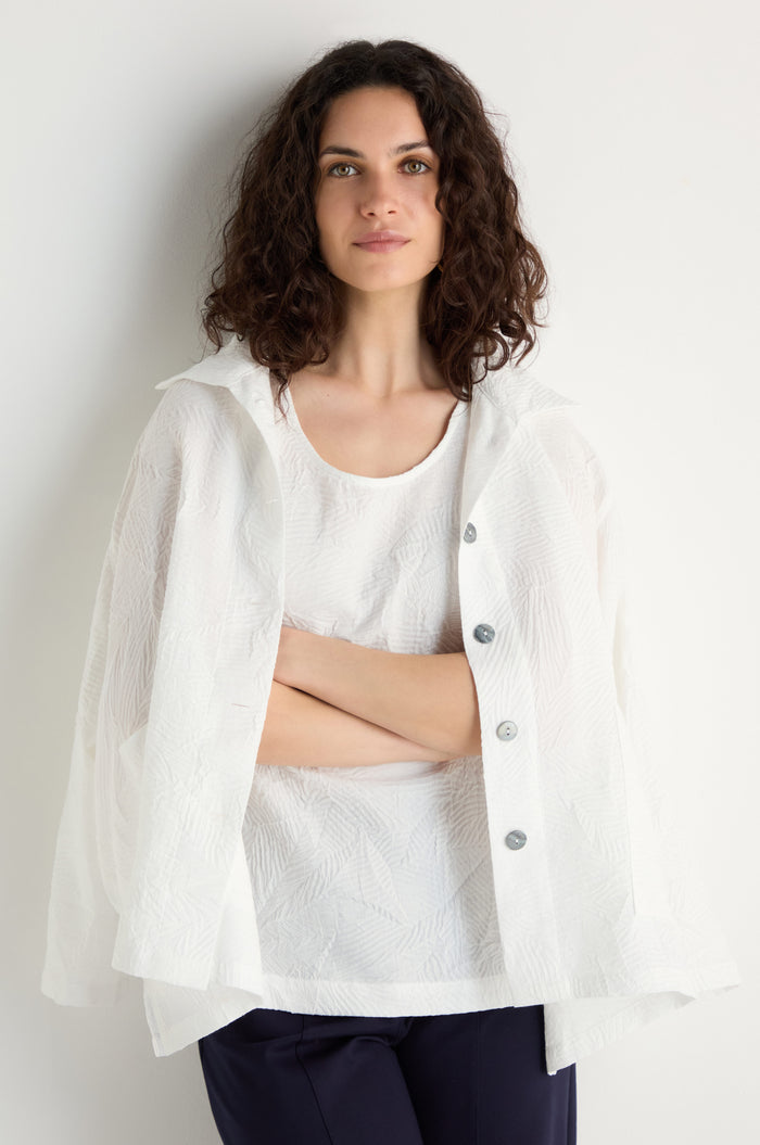Woman with wavy brown hair stands against a plain light wall, arms crossed, wearing the Pleated Leaf Top and a matching white cape featuring a raised leaf pattern.