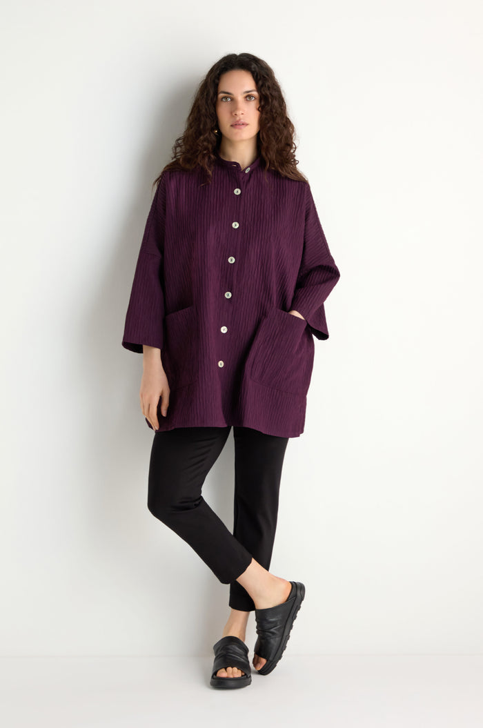 A woman poses against a white background in the Pleated Pocket Jacket, dark purple, styled with black pants and sandals. Her curly hair frames her face as she stands with a neutral expression.
