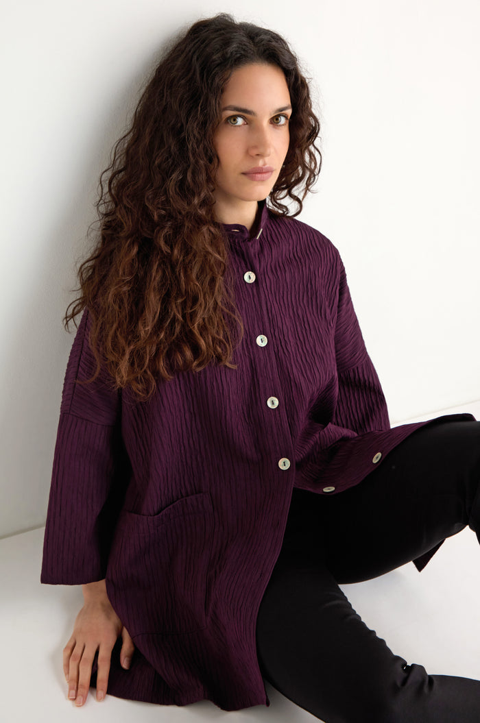 A woman with long curly hair wears the Pleated Pocket Jacket in purple with black pants, sitting against a white wall and looking at the camera.