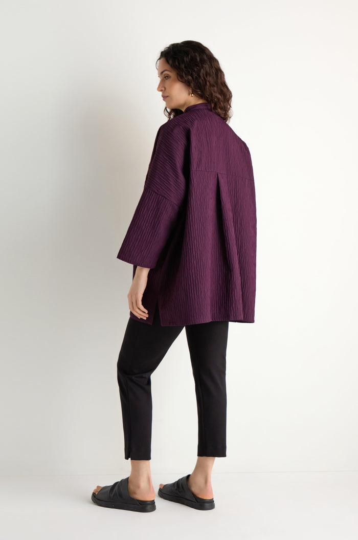 A woman stands sideways against a white background, wearing the Pleated Pocket Jacket in purple three-dimensional pleated fabric, paired with black pants and black slide sandals.