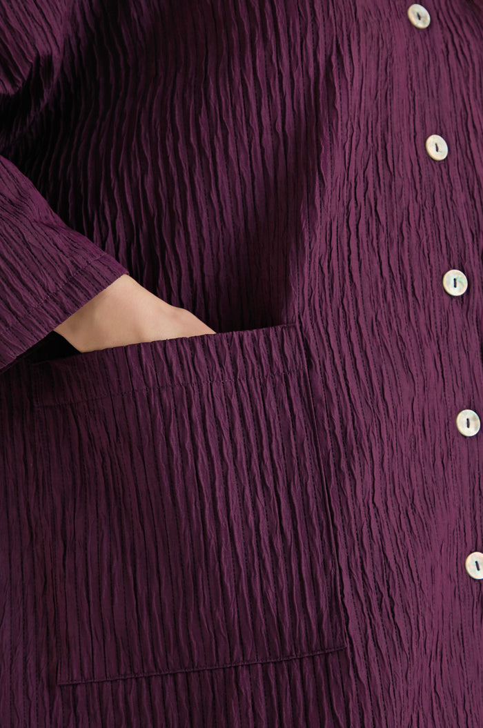 Close-up of a woman's hand in the pocket of the Pleated Pocket Jacket, featuring a plum color, white buttons, and crafted from three-dimensional pleated fabric.
