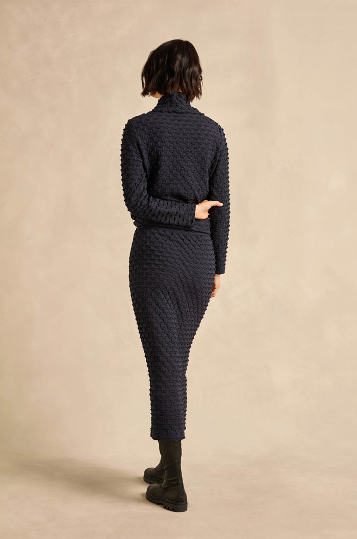 A person wearing the Pocket Jersey Tube Skirt and black boots stands facing away from the camera, with one arm bent behind their back, against a beige background.