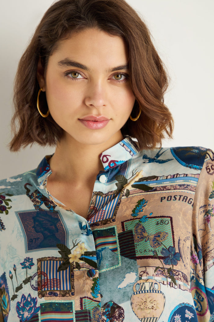 A woman with short brown hair wears gold hoop earrings and a relaxed silhouette Postcard Print Cupro Shirt with floral designs, standing against a plain light background.