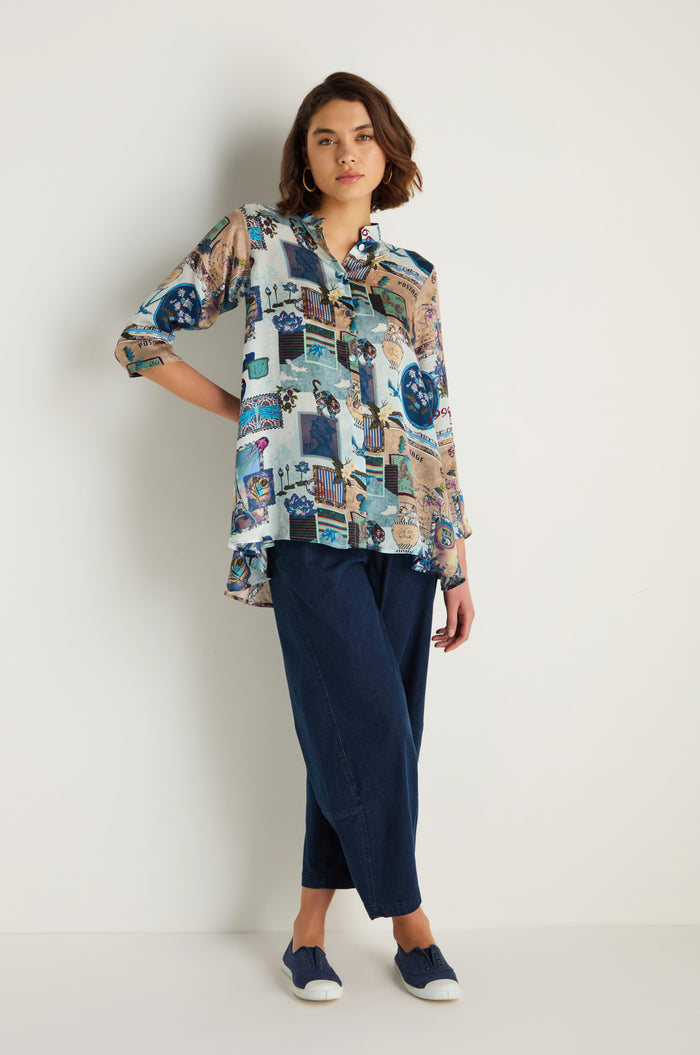 A person stands against a white wall, wearing the Postcard Print Cupro Shirt, loose blue pants that create a relaxed silhouette, and blue slip-on shoes.