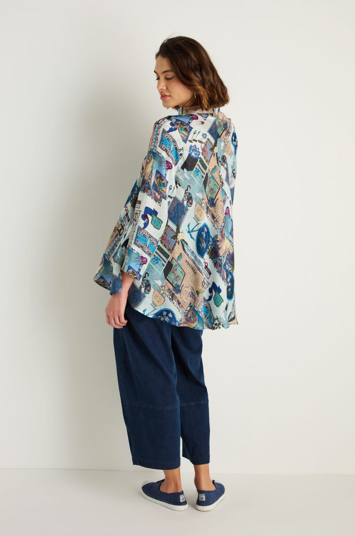 A person stands sideways in a relaxed pose, wearing the Postcard Print Cupro Shirt, wide-leg dark blue pants, and navy slip-on shoes against a plain white background.