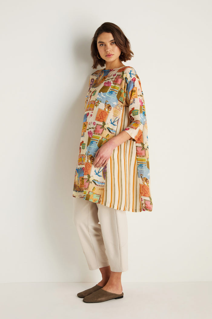 A woman stands against a white wall wearing the Postcard Print Cupro Tunic with hand-drawn prints and stripes, paired with beige cropped pants and brown flat shoes.