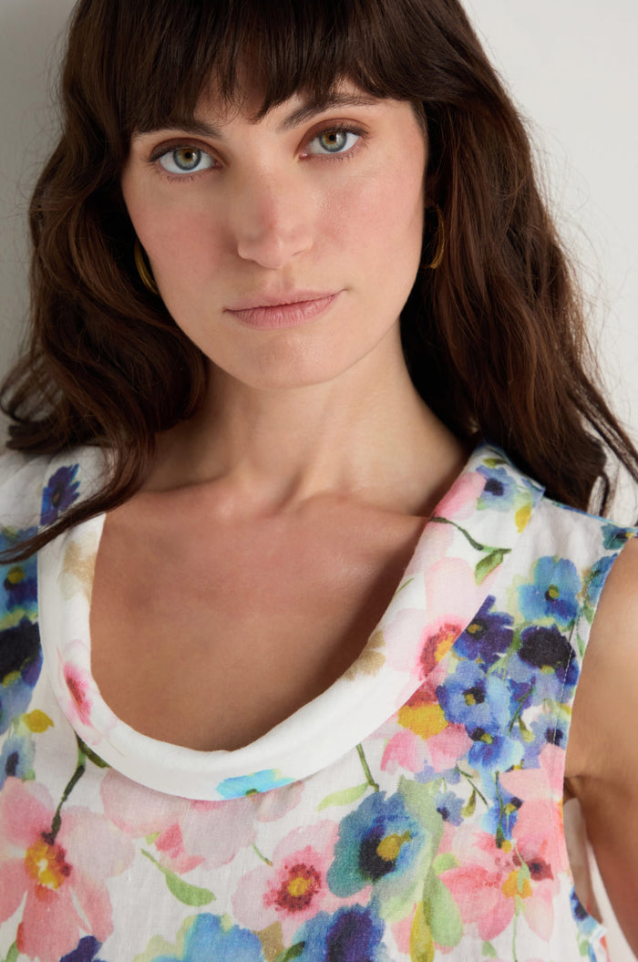 A woman with wavy brown hair models the Primrose Print Sleeveless Linen Dress, featuring a colorful floral design, as she stands against a plain background and looks at the camera.