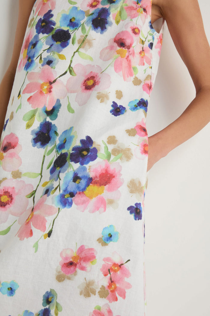 Wearing the Primrose Print Sleeveless Linen Dress, a person stands with one arm bent and a hand in the pocket, showing off its colorful print.
