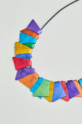 The Mini Segments Necklace features colorful anodised aluminium triangles in red, orange, yellow, green, blue, and purple on a black cord—an eye-catching piece of geometric jewelry displayed on a light background.