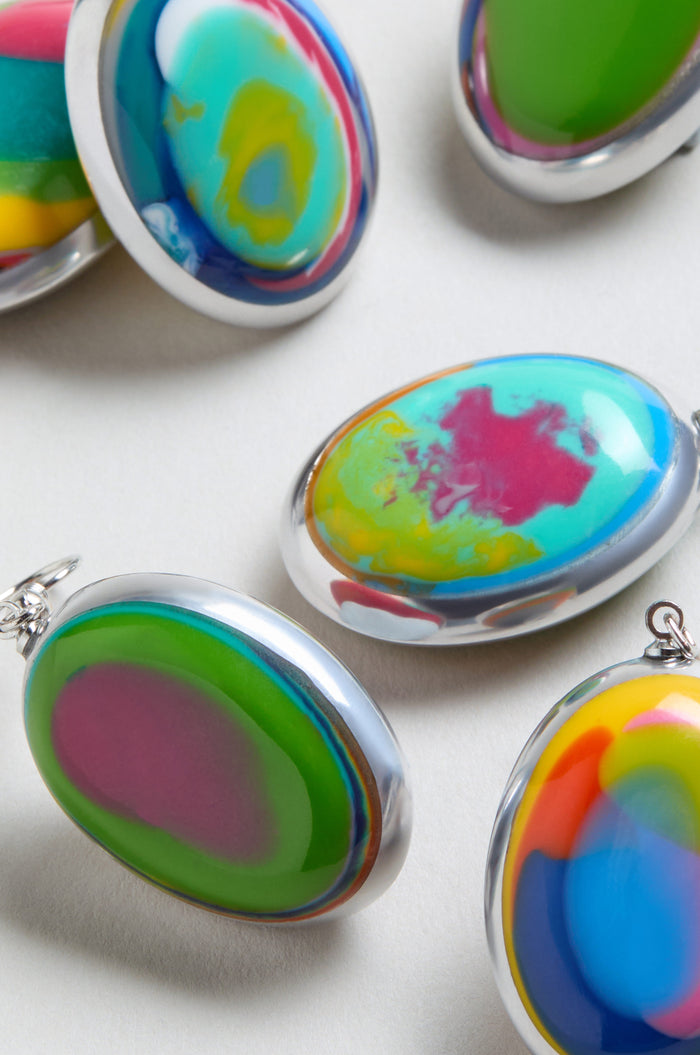 Rainbow Pebble Earrings are oval-shaped with silver edges, featuring vibrant abstract patterns. Arranged on a light surface, their multicolour design is reminiscent of resin jewelry.