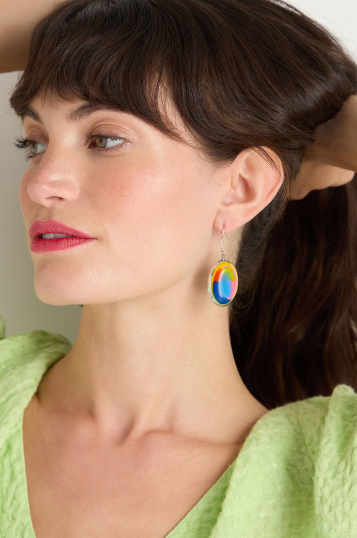 A woman with brown hair, in a green textured top and Rainbow Pebble Earrings, made from recycled aluminium and multicolour resin, looks to the side with one arm raised.