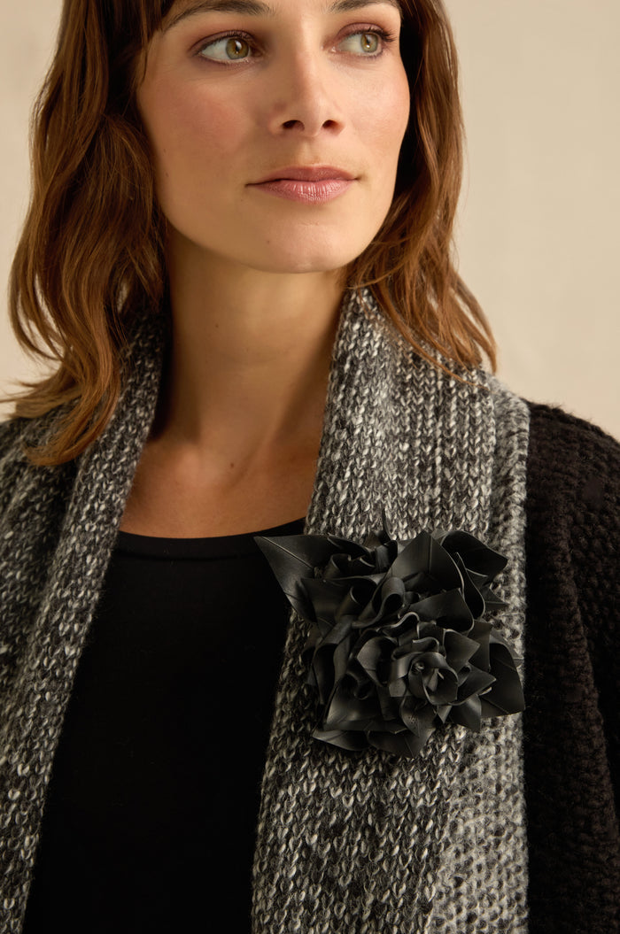 A woman gazes off to the side, wearing a textured gray scarf over a black top, adorned with the Rose Corsage—a large upcycled flower crafted from bicycle inner tube—pinned to her scarf.